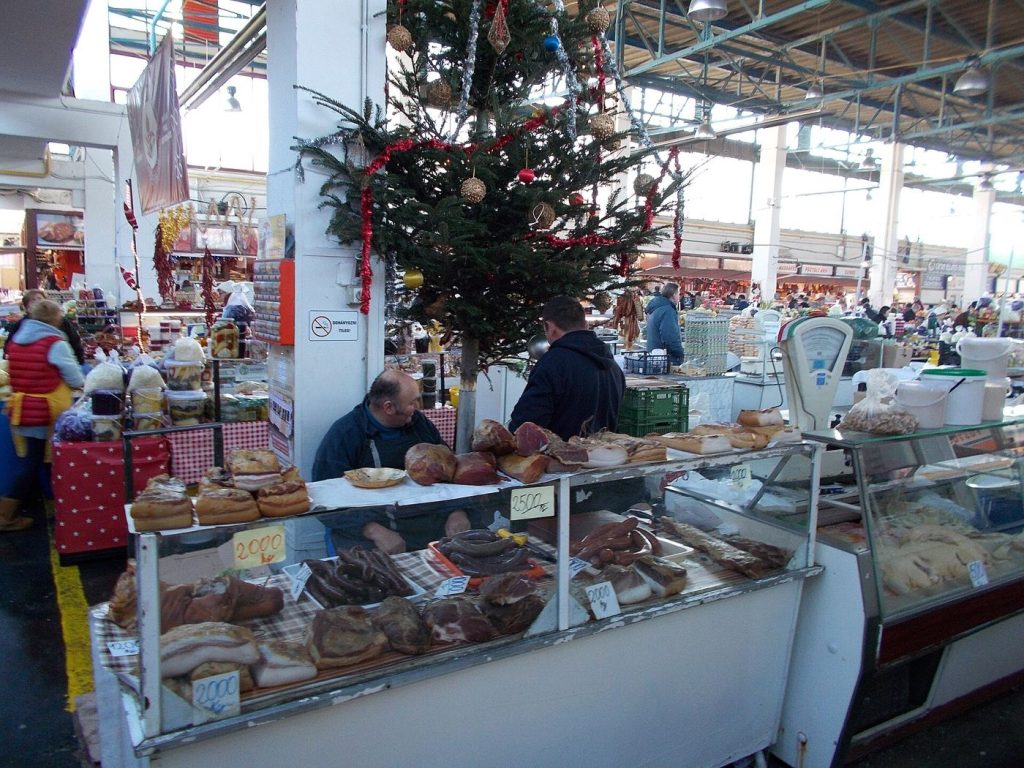 Stalls inside a bright indoor market hall selling fresh produce, bread, and flowers.